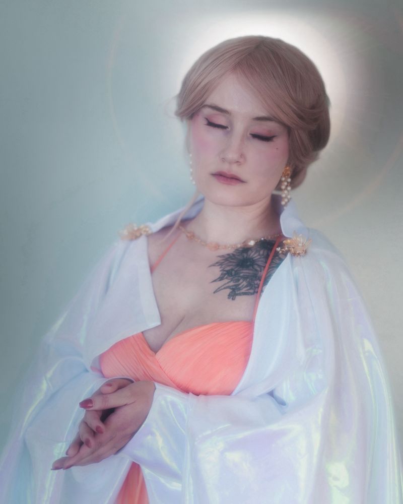 Ridley as Mercymorn from the locked tomb series. They are wearing an apricot gown and iridescent white robe, with gold/pearl jewelry. They are facing the camera with their eyes closed and their hands clasped in prayer. 