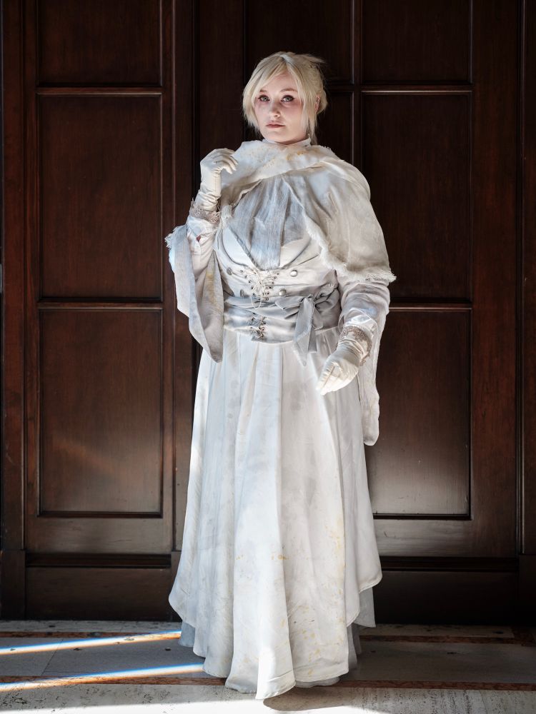 Ridley cosplaying as Iosefka from Bloodborne. They are wearing an all white garb that’s stained and weathered. They standing in front of dark wooden panels and are looking directly at the camera.