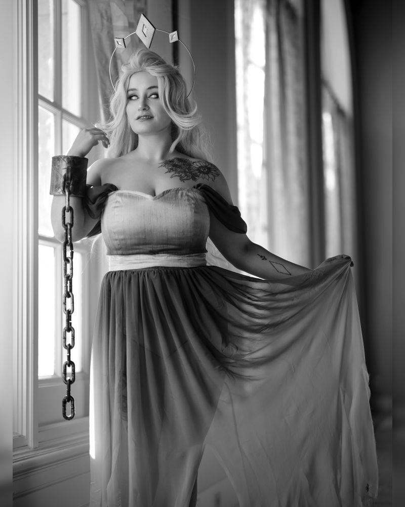 Ridley cosplaying The Tower from Slay the Princess. They are wearing a sleeveless gown with a semi transparent long skirt. The dress is a grey gradient. They are looking at the camera. 