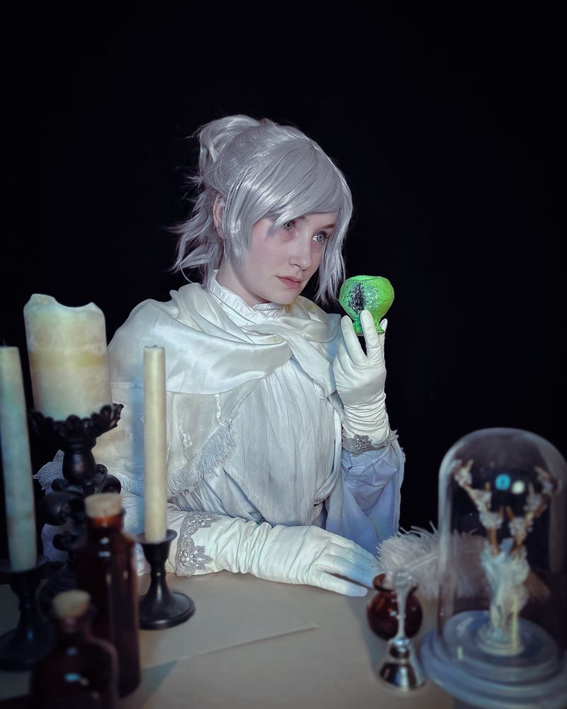 Ridley cosplaying “Iosefka” from Bloodborne. Half body portrait, sitting at a table with candles, glass bottles and various knickknacks. They are wearing all white that is distressed and stained with the grime of Yharnam. They are looking off to the side, while holding the Isz Chalice in their left hand