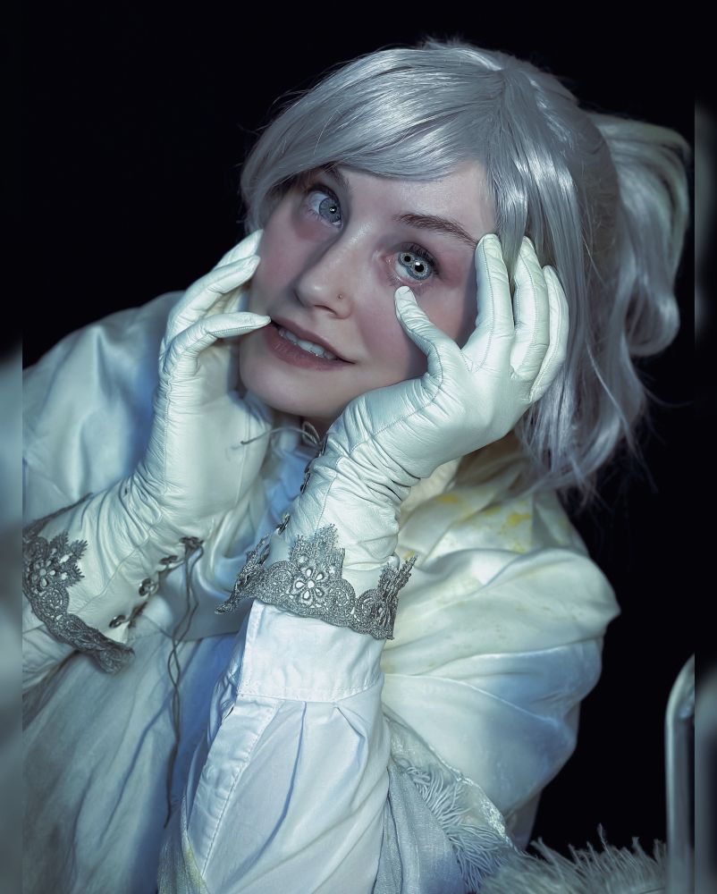 Ridley cosplaying “Iosefka” from Bloodborne. They are looking at the camera, smiling. Their left eye depicts 2 irises in mid transformation into something greater. 