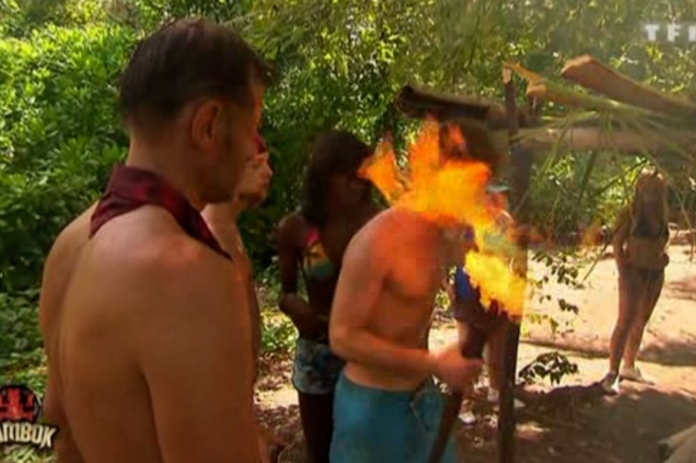 print de koh-lanta onde alguns participantes, 3 homens e duas mulheres, estão reunidos ao lado da cabana deles enquanto um participante segura uma tocha que inflama muito e o fogo atinge sua cara. As pessoas estão no meio de uma floresta, os homens sem camisa e as mulheres de biqueine, vemos o logotipo do programa em laranja no canto direito da tela e mambok escrito embaixo, o nome da equipe