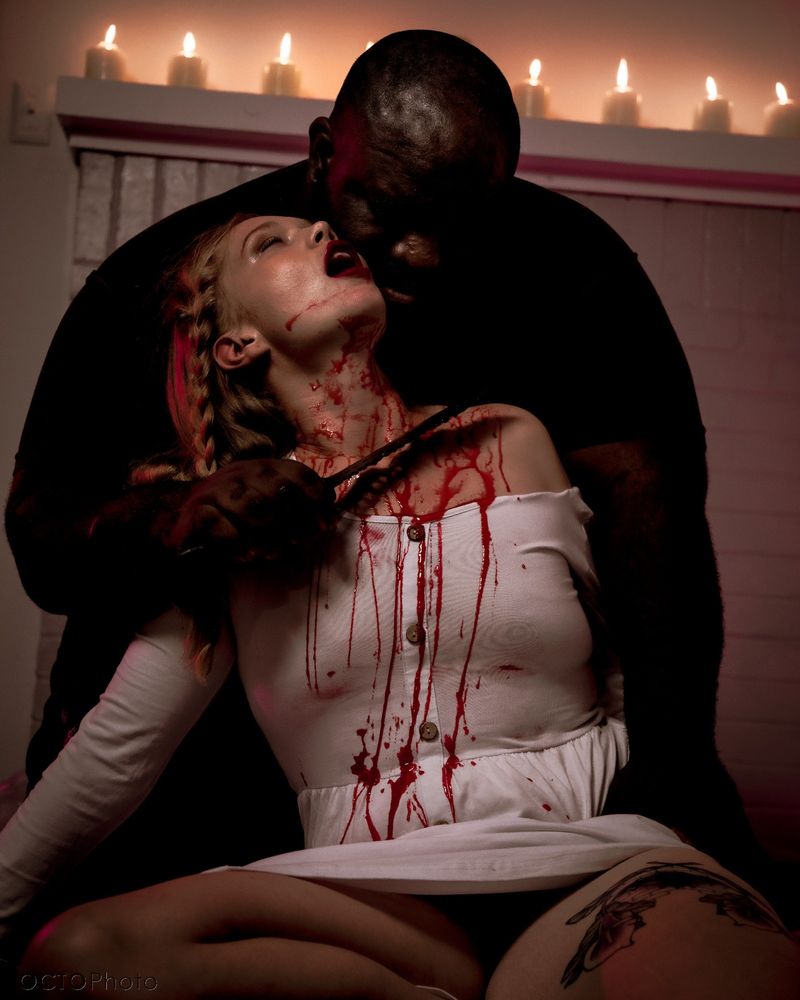 A large dark masculine figure looms behind a lighter femme figure, holding a knife close to their neck as fake blood runs down the front of them