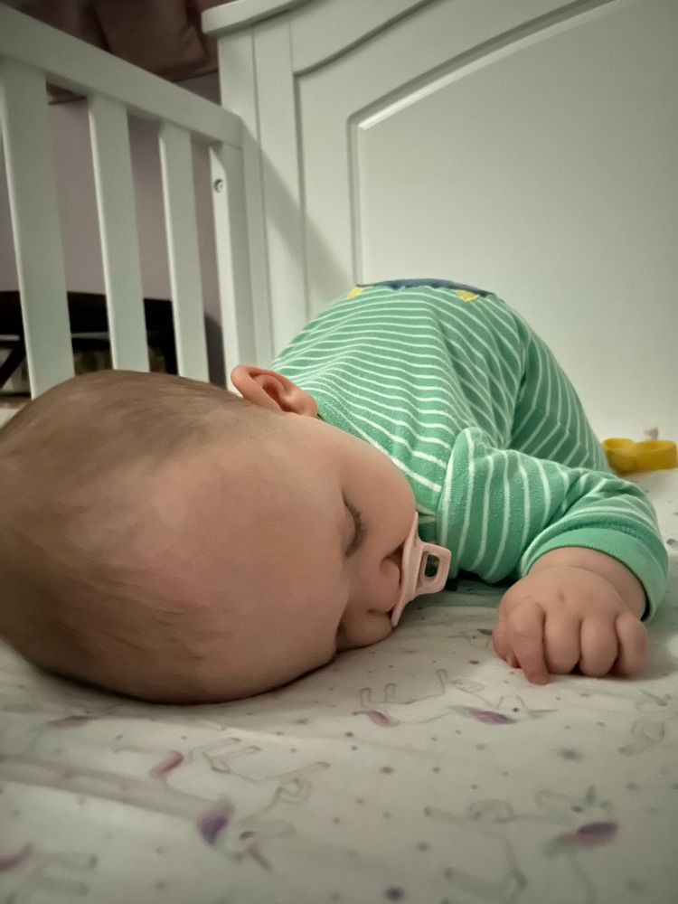 photo of my daughter asleep in the crib

she’s in a green and white striped sleep suit and lying on her tummy with her head in the foreground and her lil butt poking up

she’s got a pink pacifier in her mouth and her left hand is curled up in front of her face

she’s perfect shut up