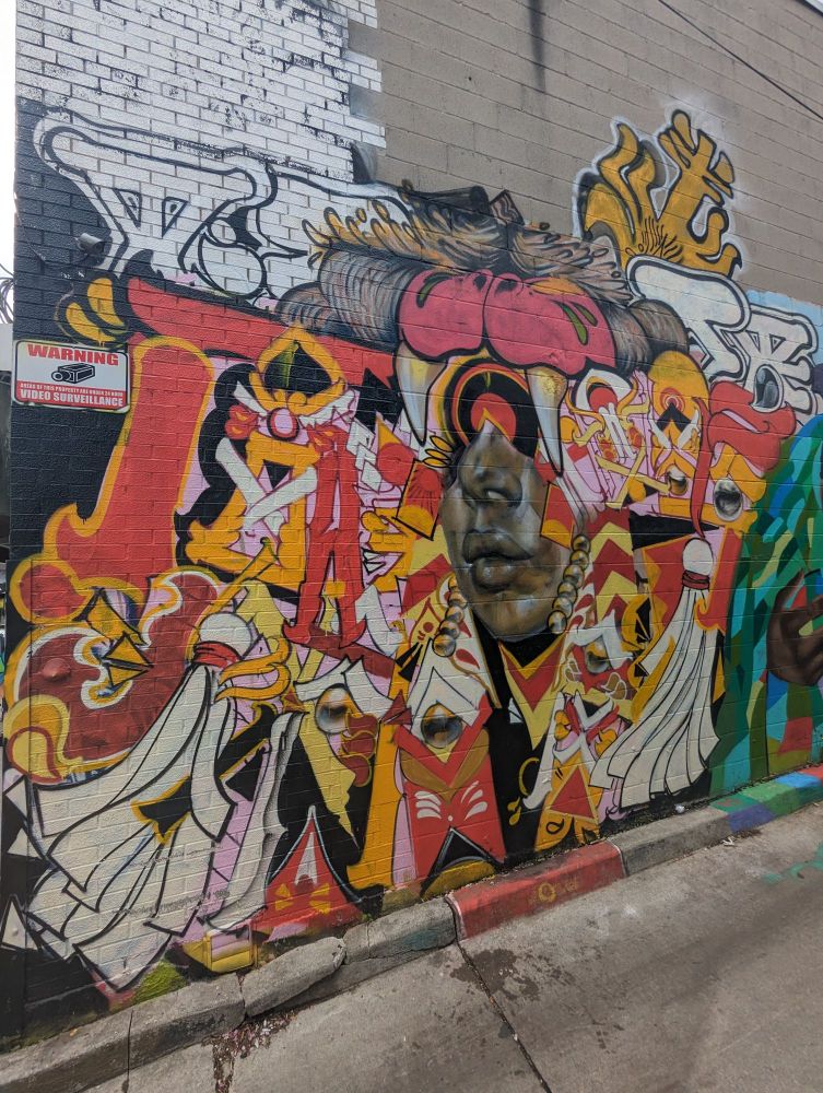 A mix of mythic symbols in red orange white brown and pink surrounding a photo realistic lower half of a man's face. A style described as "Ancient Graffuturism"