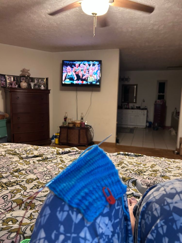 Beginning of blue all on knotting needles with Tv showing basketball in background