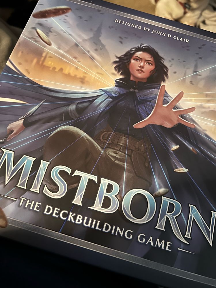 Box cover for Mistborn: The Deckbuilding Game from Brotherwise Games. A depiction of the character, Vin, from the Mistborn book series “pushing” coins away from her, presumably attacking an unseen enemy. 