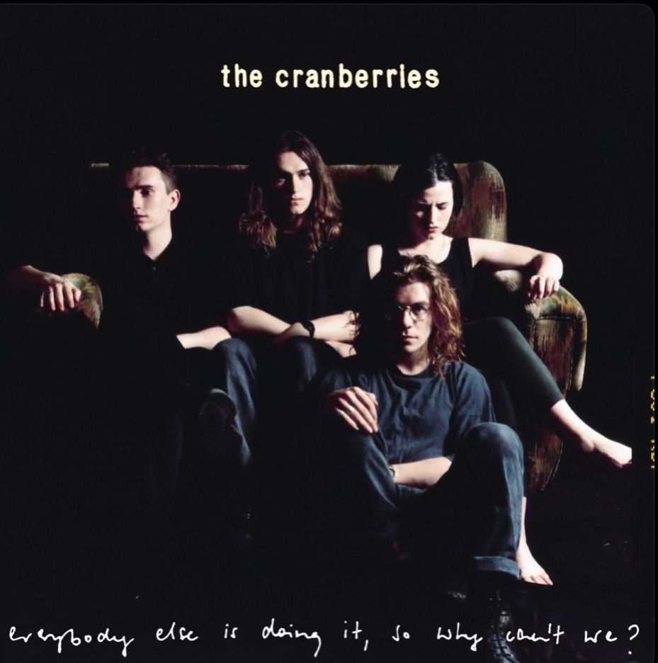 The Cranberries “everybody else is doing it, so why can’t we?” album cover 
three band members are on a couch and one on the floor against a black background. at the top “the cranberries” and at the bottom the words “everybody else is doing it, so why can’t we?” are written in cursive letters 