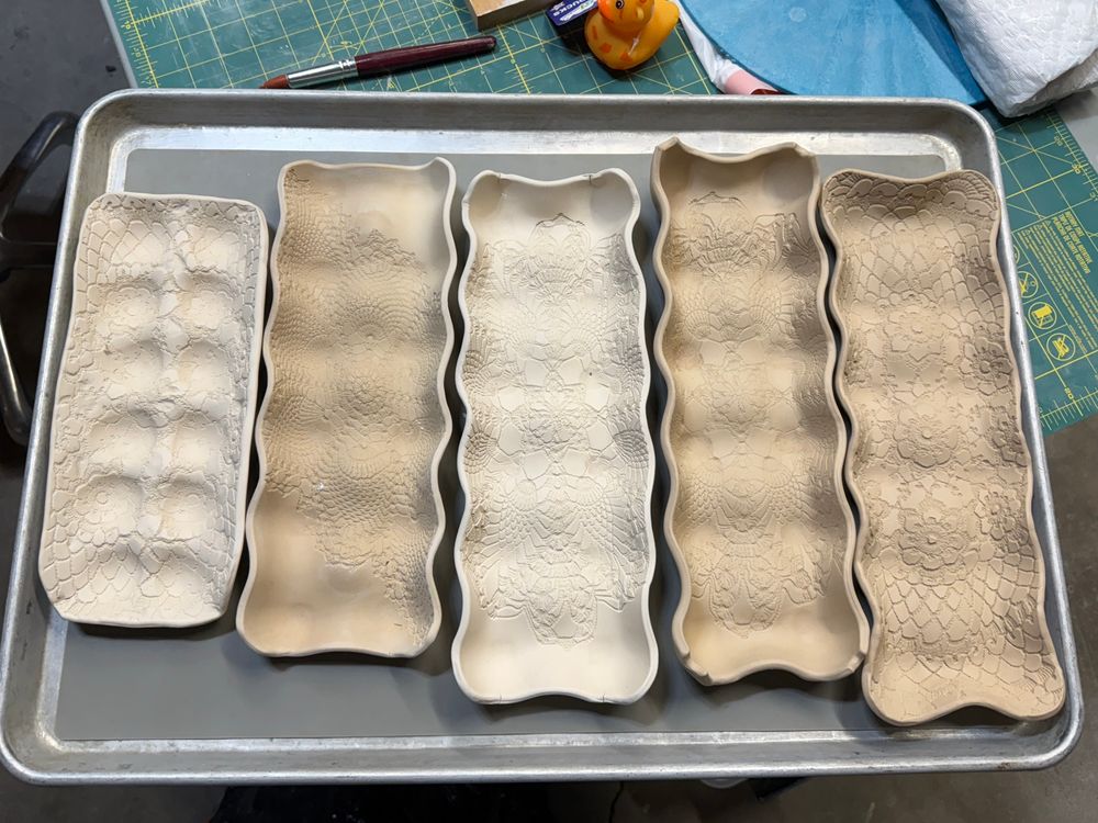 Five egg trays made of clay, all with crochet patterns imprinted into the clay. They are presented from first attempt on the left to most recent creation on the right, all resting on a full sized sheet pan. The clay is in various stages of drying. 