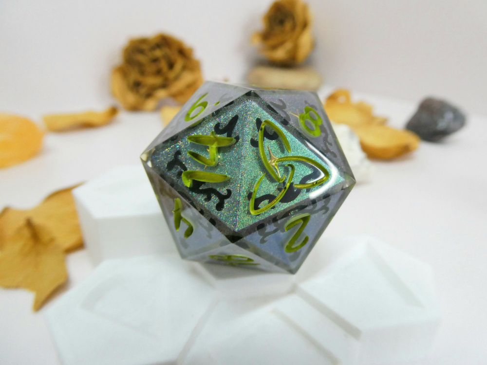 An oversized d20 with a green holographic color-shifting blank that has art nouveau styled vine details painted black on it. There are gold 4 pointed stars on the opposite vertices of the design. The numbers are painted a bright color-shift green to gold. It's sitting on a white display with yellow petals in the background.