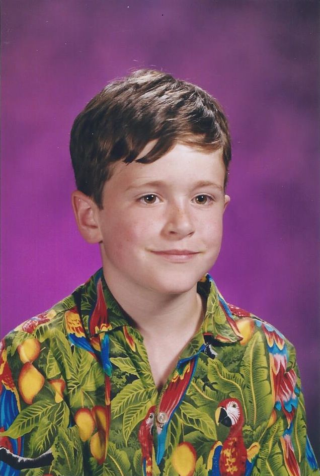 Me young, in a mostly green aloha shirt with red parrots. My smile is slightly smirking, hair is dark brown, I'm very white, background is cloudy purple. Baller school photo.