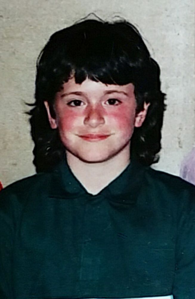 Rosy-cheeked and fair-skinned smiling boy in a dark green polo, buttoned to the top. His hair is black, long in the back, with bangs that end above his eyebrows. The photo is cropped for the privacy of others, and was taken at an opera house in San Francisco in the 90s.