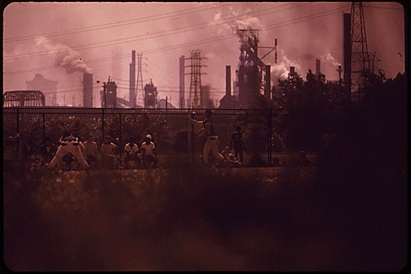 In this pink/gray photo, you can barely make out children playing baseball on Clark Fields in Cleveland, in the shadow of the Republic Steel Mill, which belches smoke into the air in the background. The photo is from EPA archives.