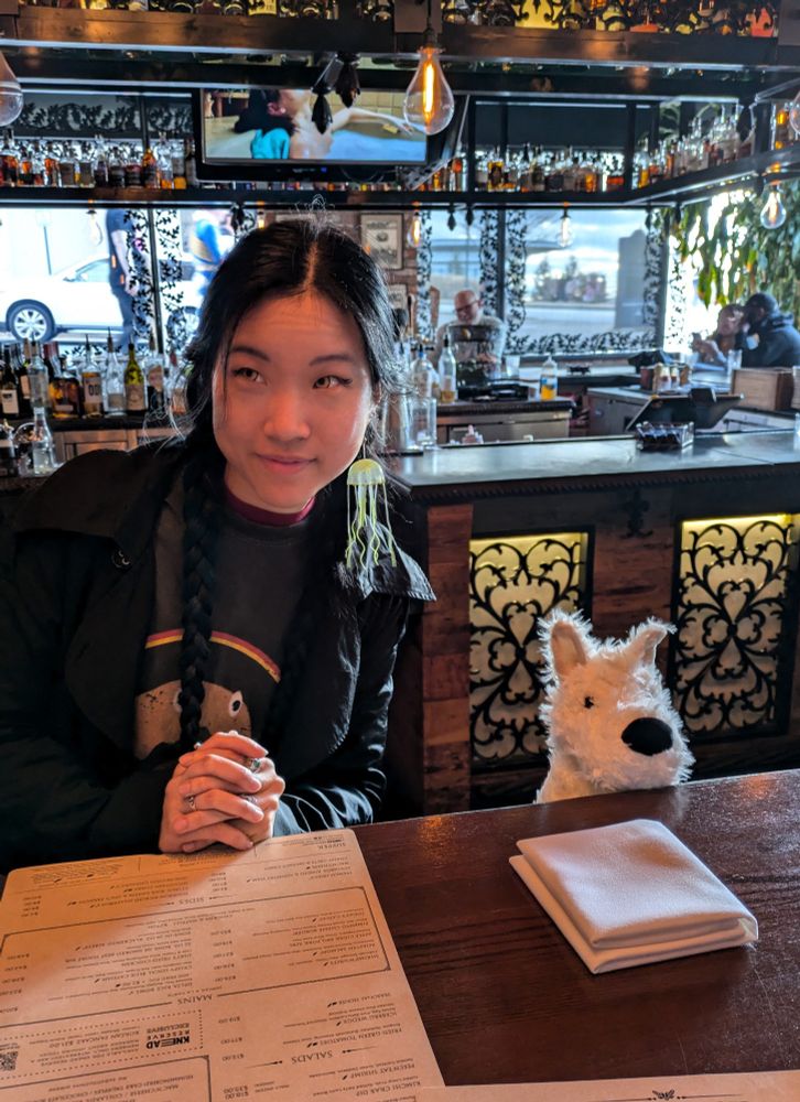 a cosplay depiction of Nona, sitting at a table at a restaurant with a white dog plushie representing Noodle seated next to her. Nona is smiling and looking off-camera.