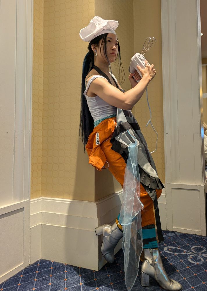 imagine Chell from Portal but a hand mixer instead of a portal gun, a companion cube apron, and a white chef's hat. she's leaning against a wall with the mixer held...like a portal gun. it's also cursed 