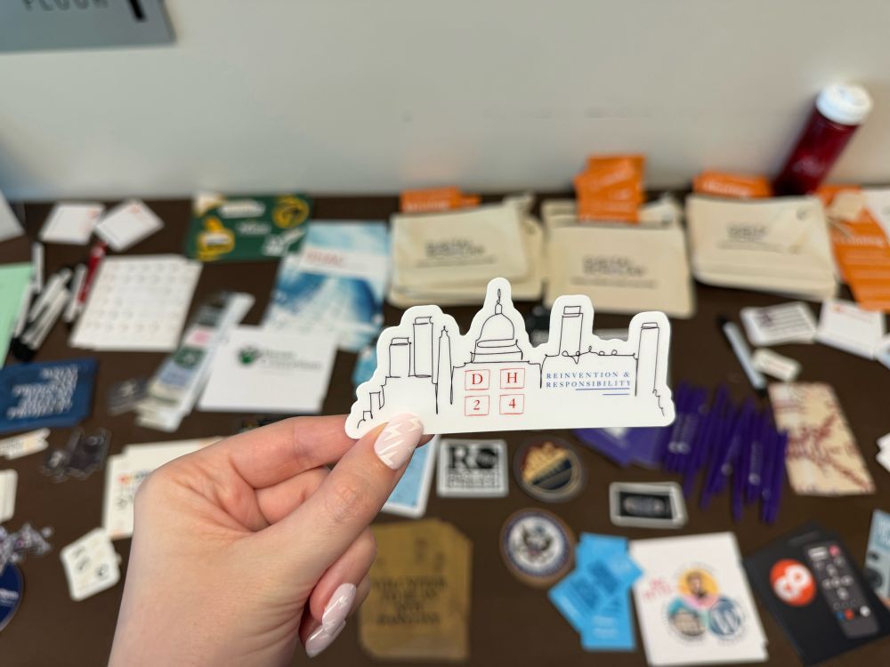 DH2024 sticker being held up with the “take swag leave swag” table in the background.