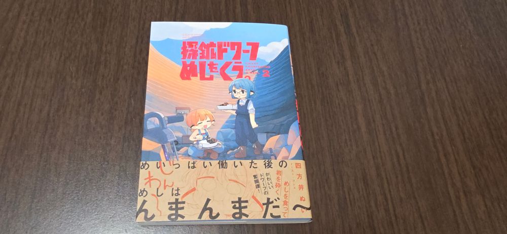 本の写真。木目調のテーブルの上に漫画の単行本が置いてある。四方井ぬい『探鉱ドワーフめしをくう。』2巻(イースト・プレス、2025年)。
