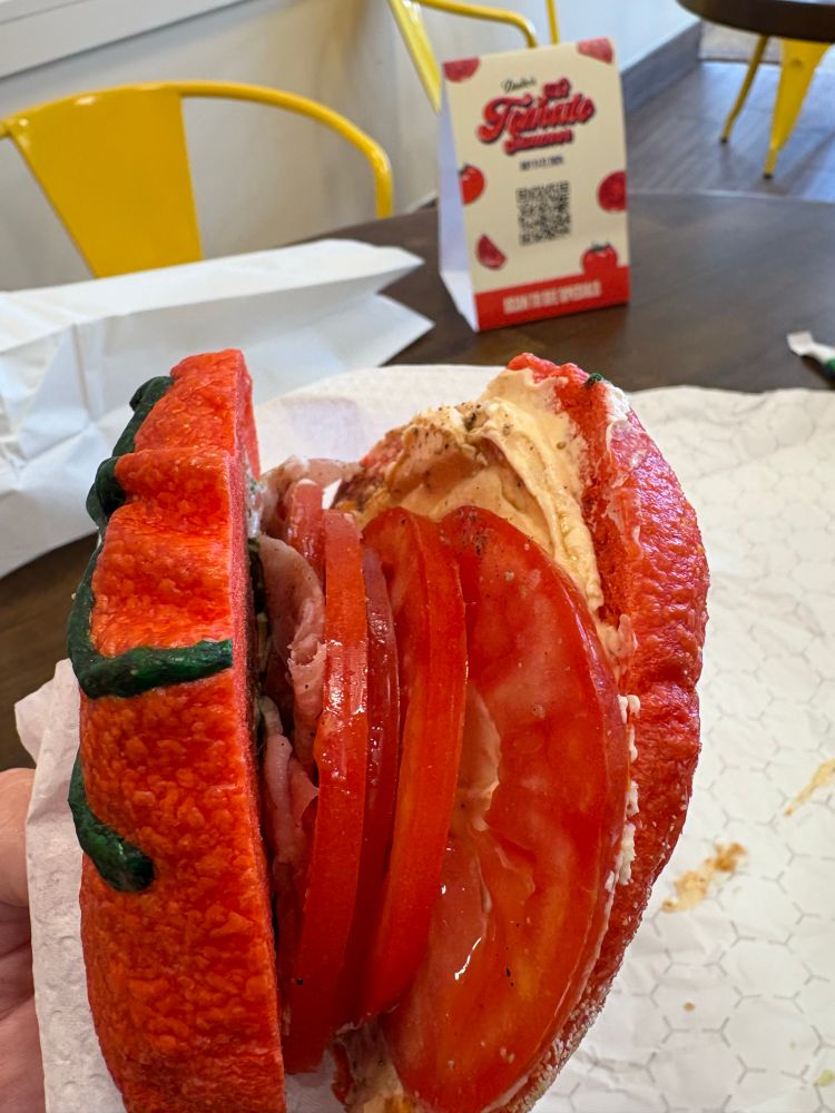 A side view of a tomato bagel sandwich in which you can see lots of tomato slices, speck (type of cured ham), and cream cheese. 
