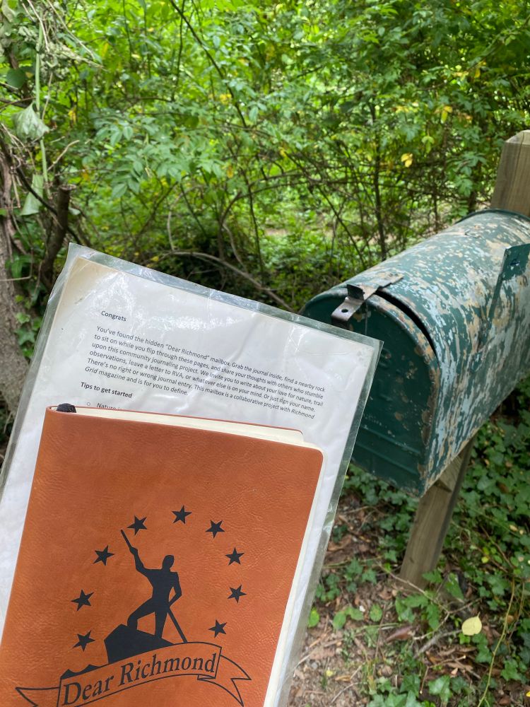An old weathered metal mailbox is tucked in the forest. In front of it someone is holding instructions and a journal that says “Dear Richmond” on the front with a silhouette of a person holding a stick and standing on a cliff or something.