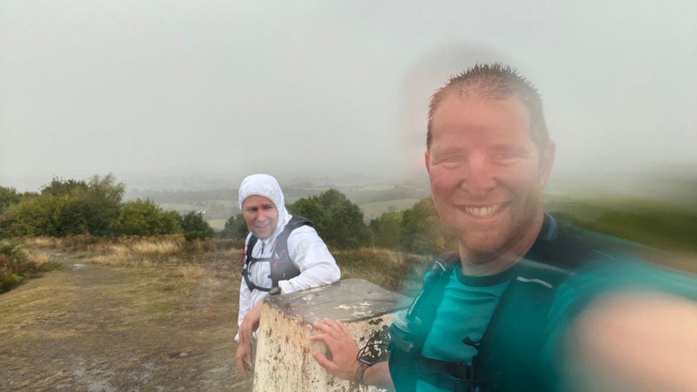 The same two men, on top of another hill. The camera lens is covered in rain. They are leaning on a trig point. They are drenched. One is smiling slightly manically. The other wears the grimace of a man who knows he’s got 20km to go and would, right now, welcome a quick, warm demise at the hands of the approaching thunderstorm.