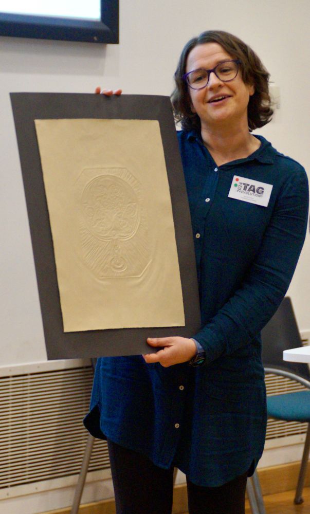 Photograph of a woman with dark wavy hair, spectacles, and a dark green long-sleeve dress, holding a piece of artwork by Alice Clough: a paper embossing of a design inspired by an Iron Age mirror.