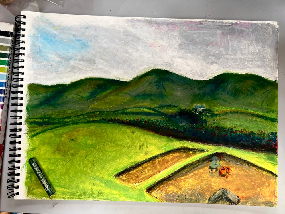 Photograph of an A3 sketchbook page with a rough ‘mood’ sketch in oil pastels, showing an archaeological excavation of one of several undulations in a pasture field. A single archaeologist fills orange buckets. In the background, the land rises to a range of hills.