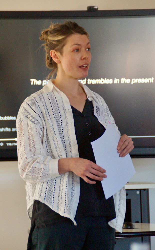 Photograph of a woman with fair hair and a white shirt over dark top and trousers, holding a sheet of paper, delivering a conference paper in front of a screen bearing the words “the past fizzes and trembles in the present”.