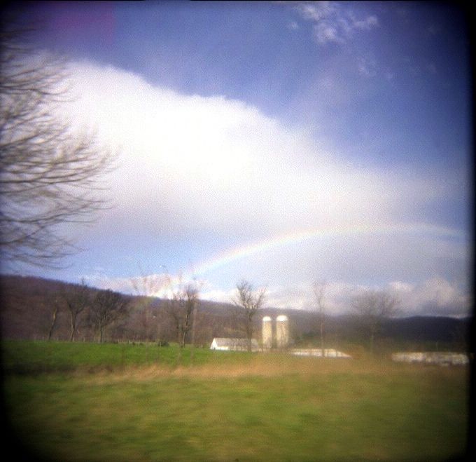 Grass, blue sky with a big white cloud and a rainbow arching over two silos on a farm. The square frame and a black vignette signal this was taken with a Holga camera.