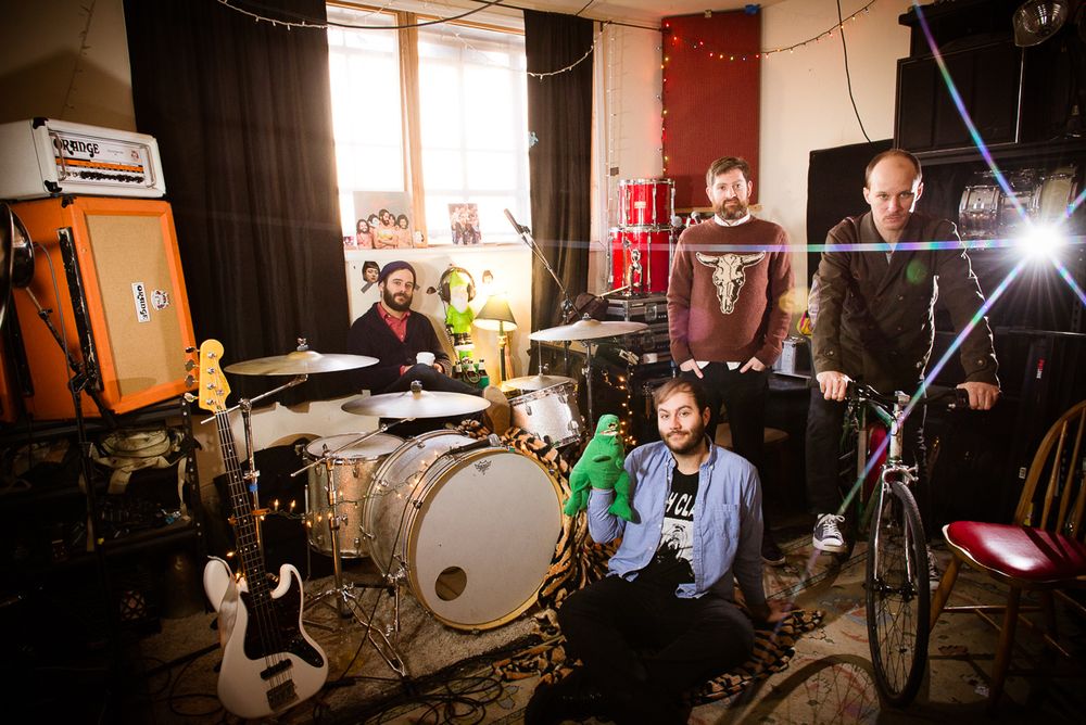 A promo shot of the band MiniBoone of four guys in a band practice space. One on a bicyle, one holding a pocket, one behind a drumset, one standing with his hands in his pockets.