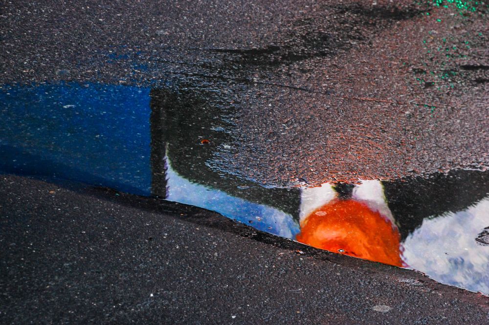 The pavement with a puddle reflecting an upside down part of a man’s face wearing a tie and suit.