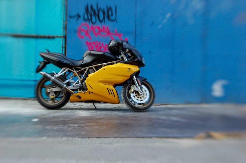 A yellow motorcycle with a blue wall behind tagged with pink/black graffiti 