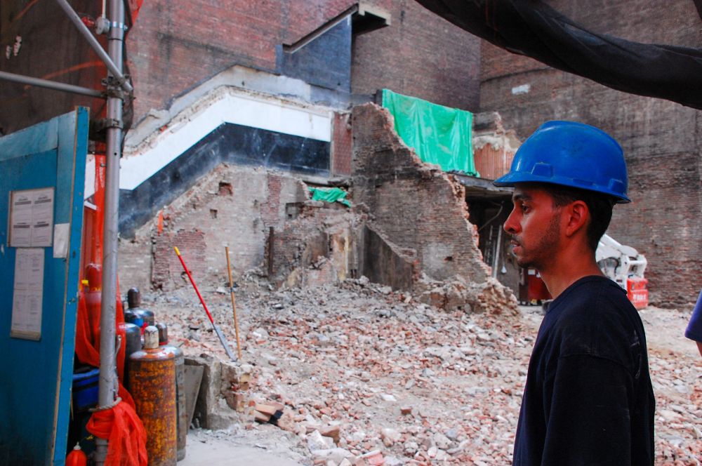 The profile of a man in a blue construction hat in front of a building demolition site 