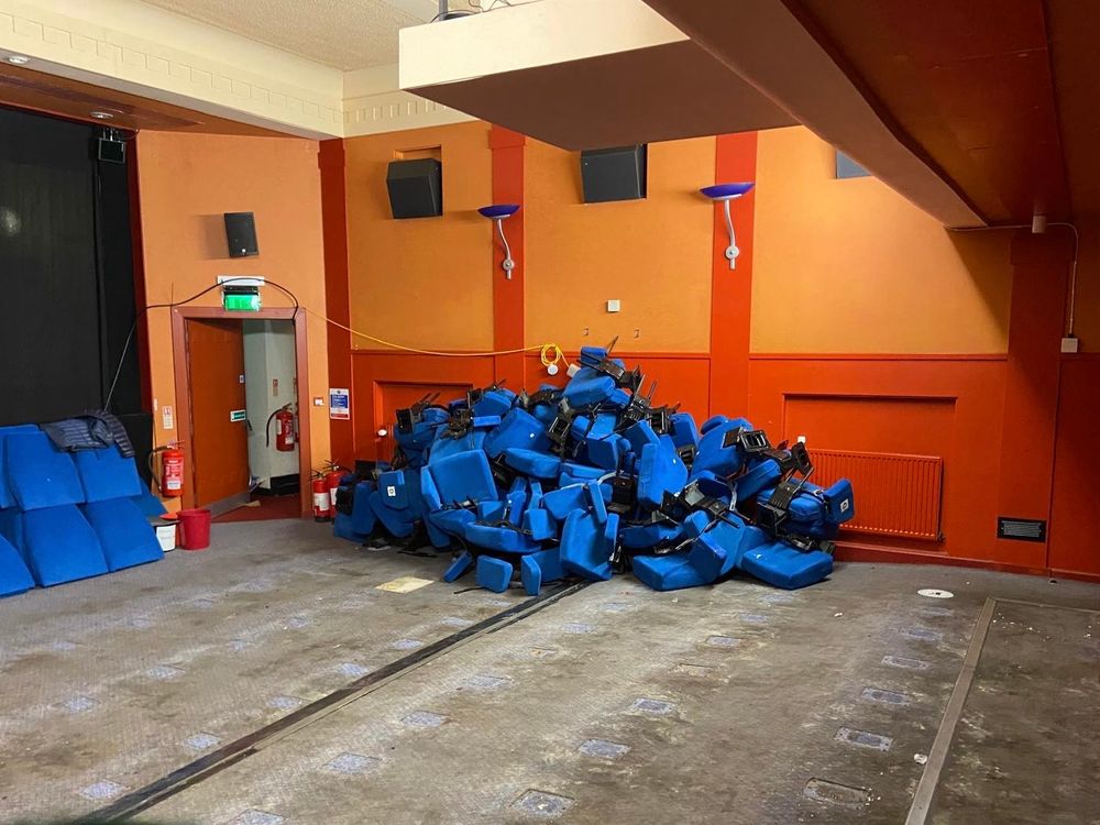 An empty cinema auditorium with a bare concrete floor and dismantled blue seats piled up against the wall. 