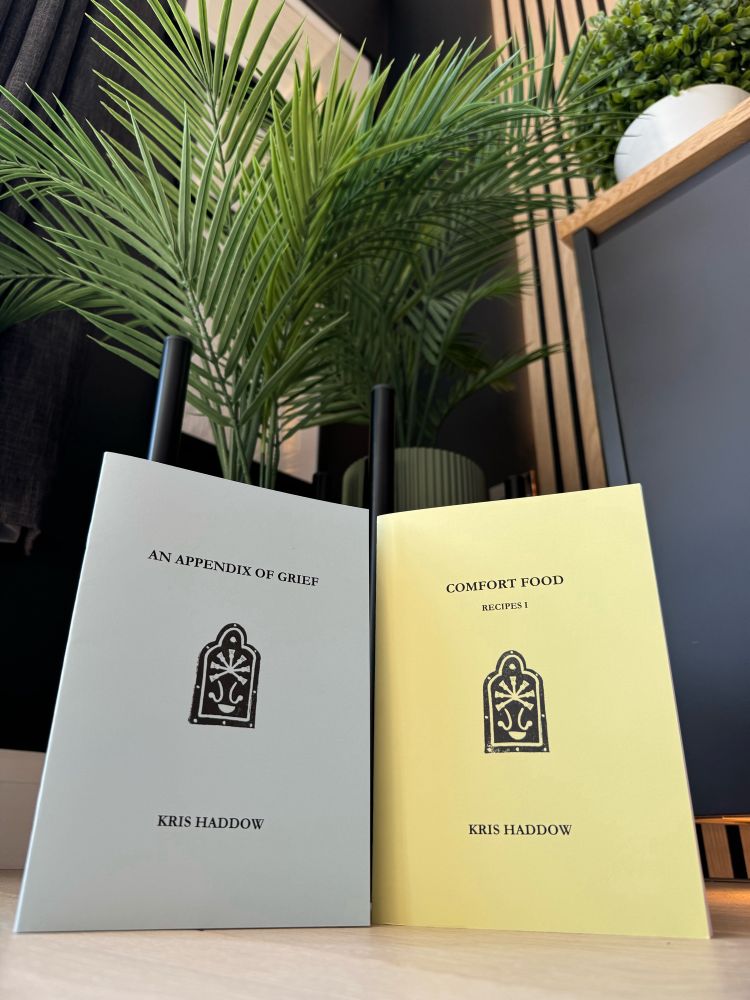 A blue booklet and a yellow booklet stand side by side under some plants. One is titled An Appendix of Grief, the other Comfort Food: Recipes I. Both are by Kris Haddow.