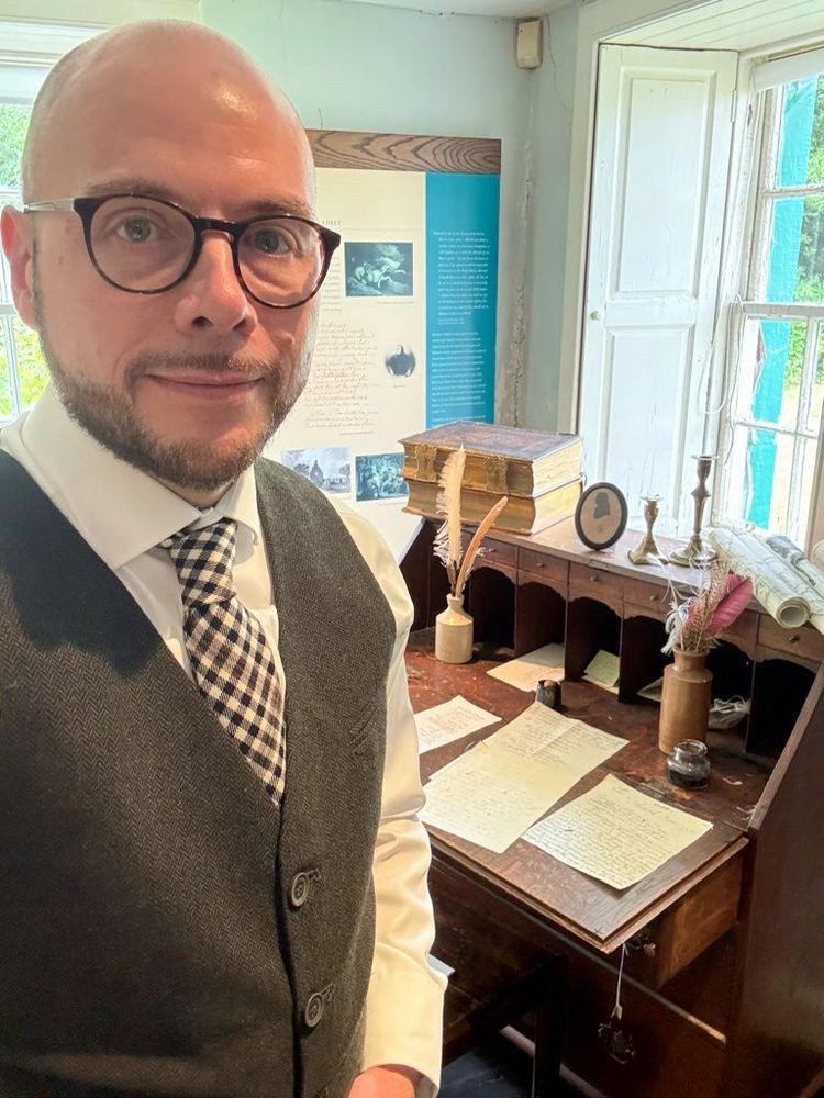 A man with a beard and round glasses wearing a waistcoat and a burns check tie. He is standing by the writing desk by the Spence in Robert Burns’ Ellisland Farm, Dumfriesshire, Scotland. Papers and quill pens are in the desk