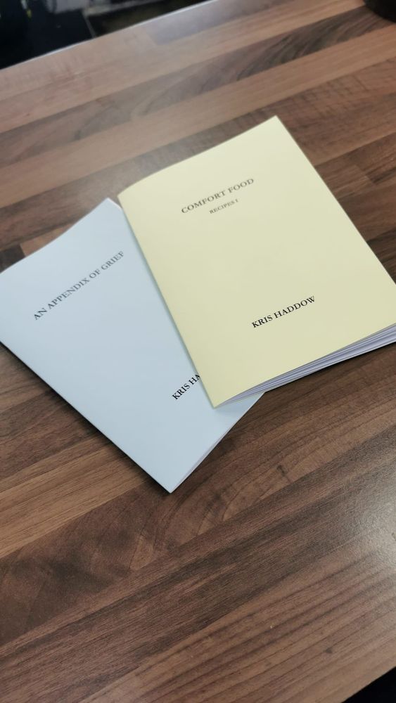 two small booklets, one light blue, the other yellow, sitting on a wooden desk. the blue one is titled an appendix of grief, the yellow comfort food. both are by kris haddow
