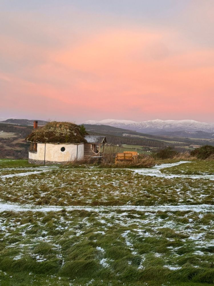 the round turf-roofed hobbit house at Moniack Mhor under a pinky-peach morning sky, snowcapped Scottish mountains in the distance