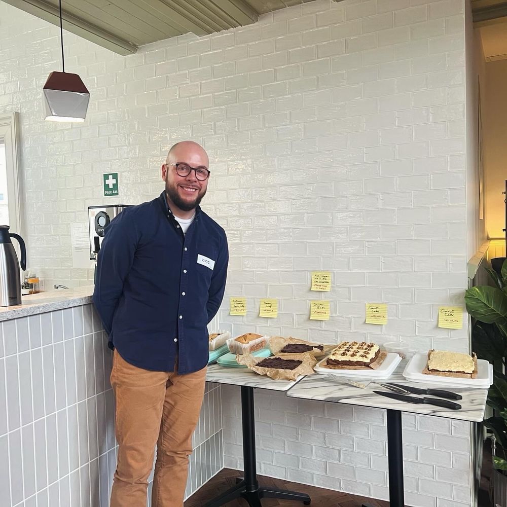 Kris, a bald man with a beard in round glasses, wearing a blue shirt and khaki trousers, standing beside a table filled with cakes