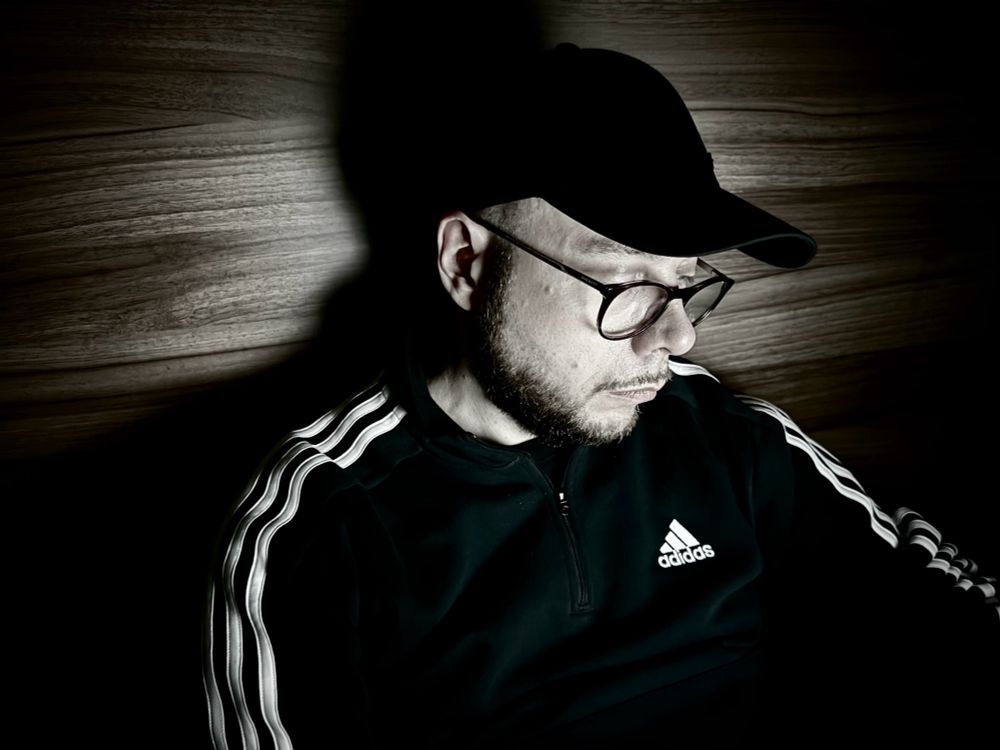 Playwright Kris Haddow wearing glasses, a baseball cap, and a black adidas top. He is coding on a laptop against a wood panel. He thinks he looks cool but he’s definitely not.