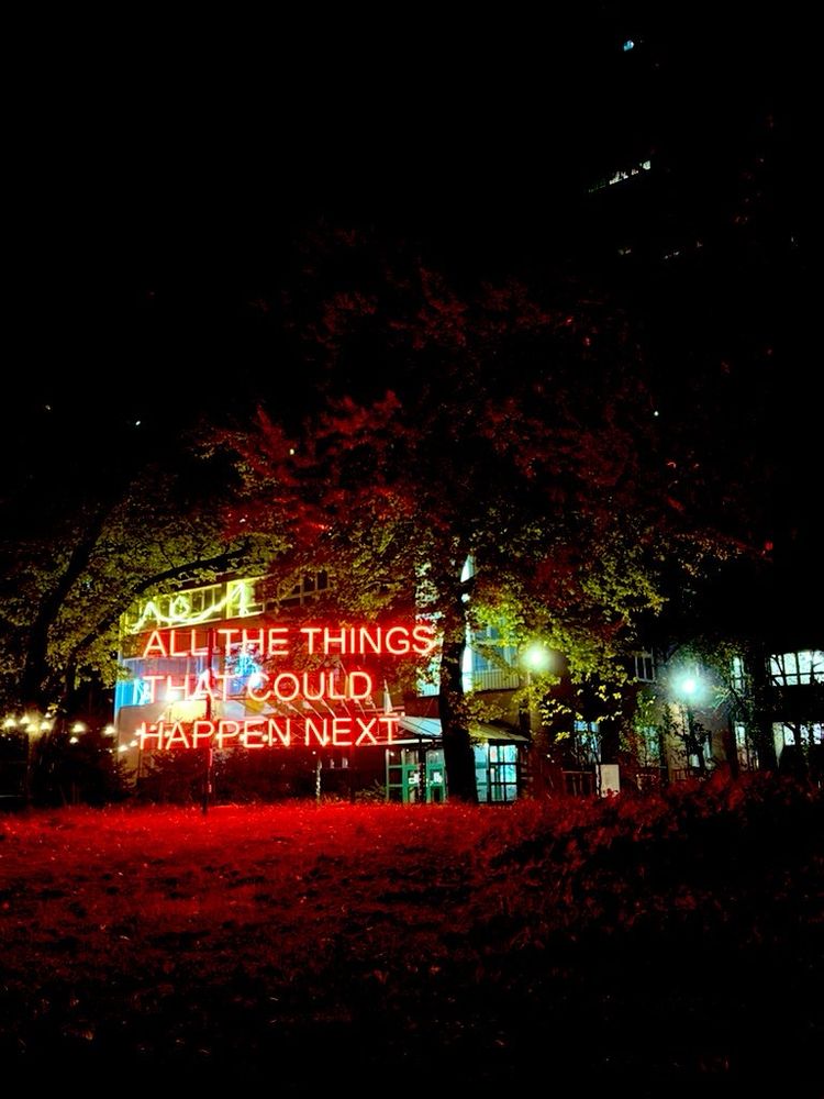 A red neon sign glowing under a tree outside a theatre in Warsaw, Poland. It says ALL THE THINGS THAT COULD HAPPEN NEXT in uppercase. The leaves on the ground are bathed red.