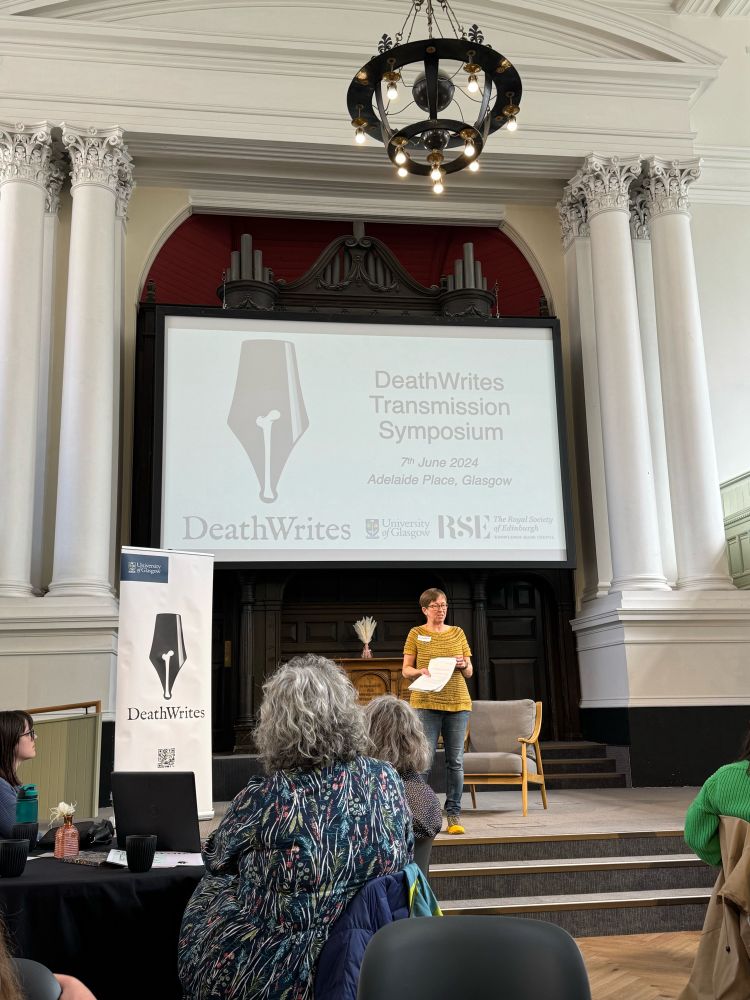 Dr Elizabeth Reeder on a stage under a large banner welcoming writers to a conference on death writing