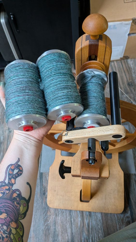 A hand holding two bobbins of blueish yarn above a spinning wheel that holds a third bobbin of the same yarn.  The spinning wheel, an old-style single treadle Schacht Matchless, is a warm honey colored wood, but the 3D printed bobbins are an incongruous shade of metallic silver with bright red gears on the end.  The yarn is roughly DK to worsted weight singles in turquoise blue with hints of purple and gray.  The person holding the two bobbins has a tattoo of a tiger kitten holding a ball of yarn on their arm.