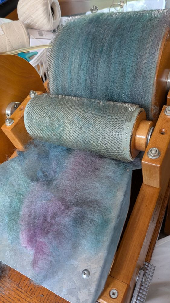 A drum carder loaded up with the turquoise and purple wool.  The wool on the drum is mostly turquoise with a few streaks of darker teal.  The feed tray has a layer of lightly fluffed wool ready to be added to the batt, which shows the various shades of lilac, blue, and sea green more clearly.  After blending, the colors will mostly mix together into a muted haze.