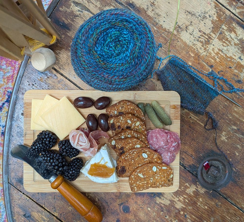 A small charcuterie board and a ball of yarn on a wooden table.  The yarn looks predominantly blue in this picture but still has striations of other dark colors.  The board has an appealing array of crackers, cheese, and cured meats, with a few blackberries and chocolate covered almonds for dessert.  It's just enough for a meal for one person, or a snack for two.