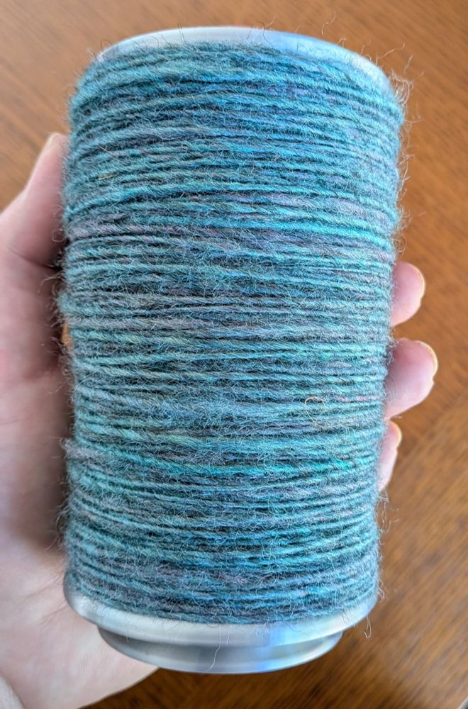 A bobbin of hand spun singles held by a person's hand.  The singles are turquoise with hints of lilac and have a woolly, fuzzy texture.