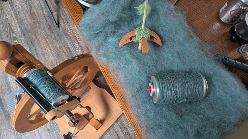 A top down view of a variety of yarn spinning paraphernalia.  On the left is a spinning wheel on the floor, bearing a nearly full bobbin of muted turquoise yarn.  On the table to the right is a spread out batt of wool, the same turquoise/gray/lavender/blue as the yarn on the spinning wheel, and there is another bobbin full of yarn sitting on the batt.  Also sitting on the batt is a Turkish spindle with a small cop of pale green yarn.