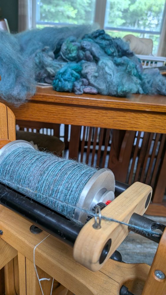 A close up view of a bobbin on a spinning wheel, mostly full of turquoise yarn.  The yarn has bits of lavender mixed in, and in the background is a pile of turquoise and purple dyed wool.