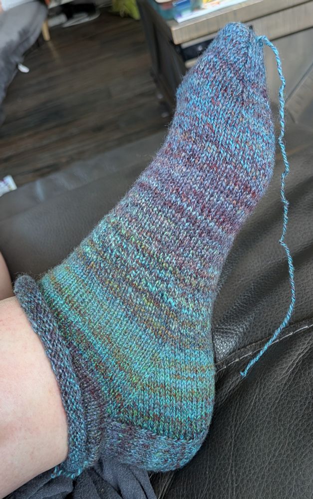 A nearly finished handspun, hand knit sock on a foot.  The sock has a short rolled cuff, and shifts in color from blue/red marls at the ankle to turquoise to ocean green to brown to purple back to bright blue again at the toe.  The only thing left to do is weave in the tail that dangles off the toe... And knit another sock, of course.