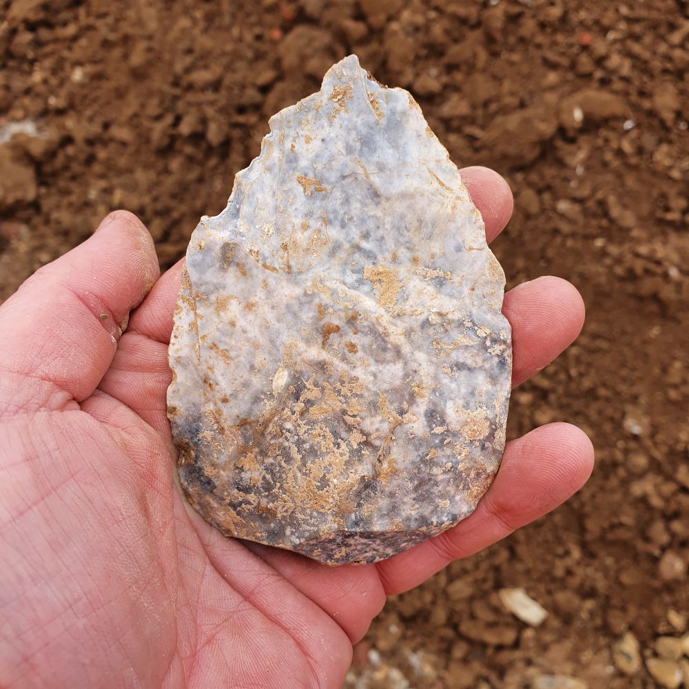 A mottled grey-white pear-shaped flint tool about 6cm across and 10cm long sitting in a hand. Gravel behind.