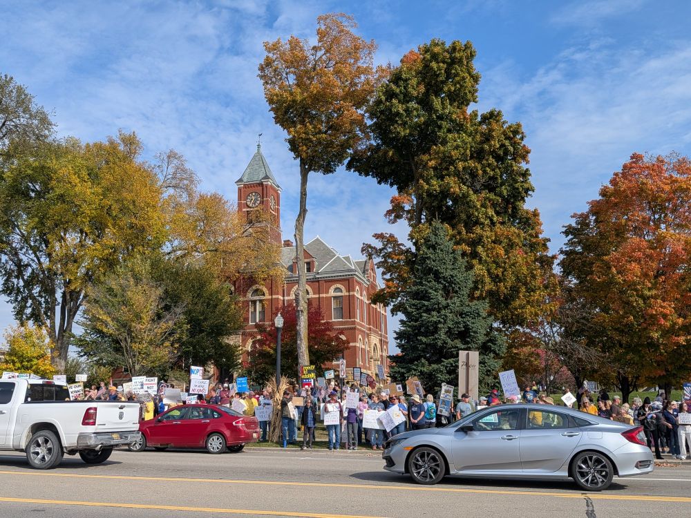 Across the street view of the historic red brick Livingston County Courthouse showing a large group of No Kings protesters holding signs as cars drive by. Blue sky and trees with autumn leaves 